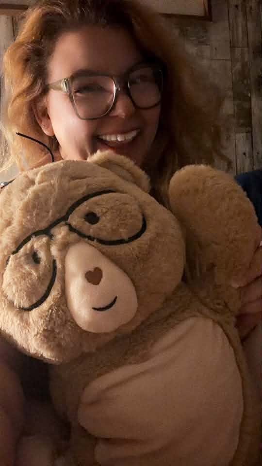 A smiling woman with curly hair and glasses holds a large plush teddy bear wearing round glasses. The photo is taken indoors with warm lighting against a rustic wooden wall.