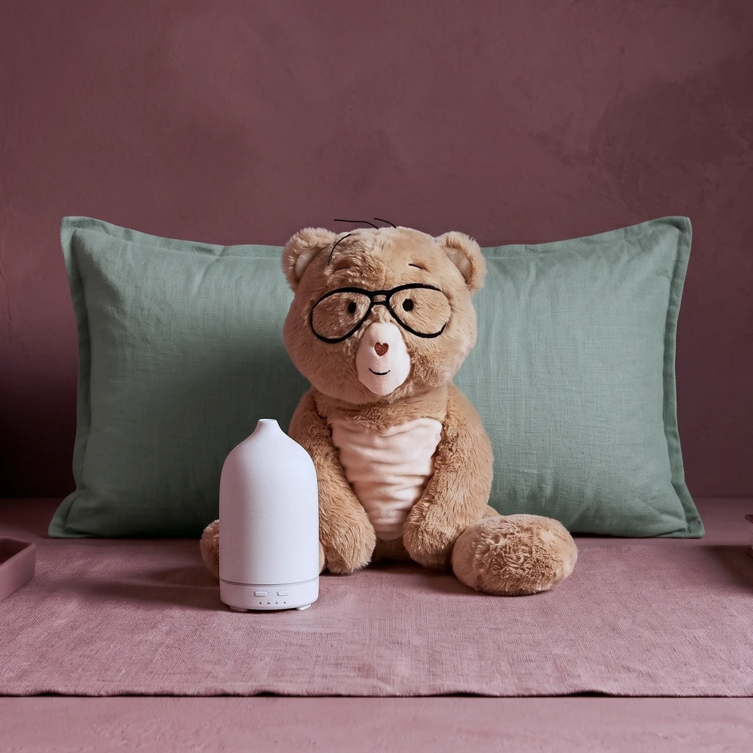 A plush teddy bear wearing black glasses and a cream shirt sits in front of a sage green pillow on a dusty rose bedspread, with a white diffuser in the foreground.