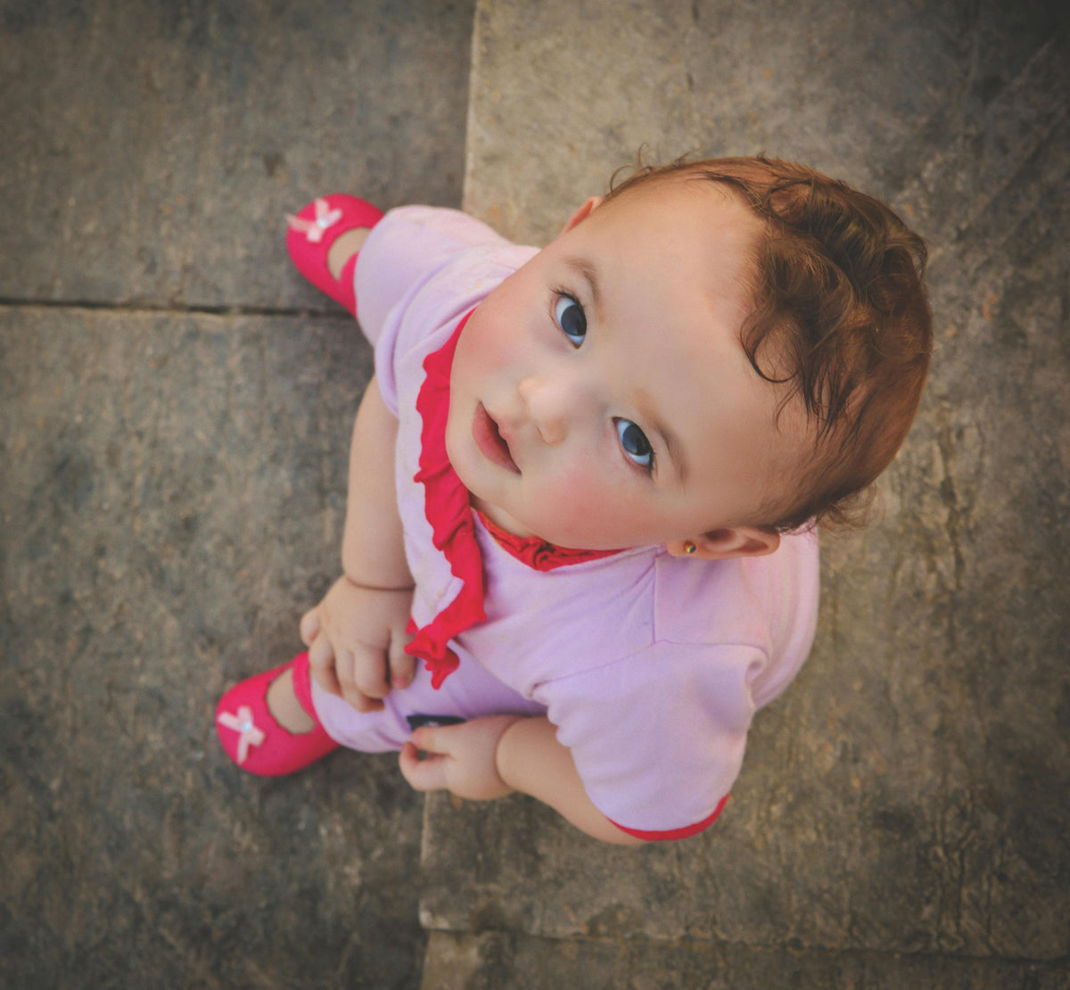 Toddler looking up at the camera, wearing pink dress with ruffles and pink shoes with bows.