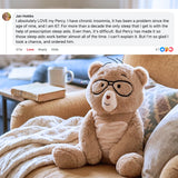 A plush teddy bear with black glasses and a cream belly patch sits on a beige couch in a living room setting, with a customer review text overlay at the top.