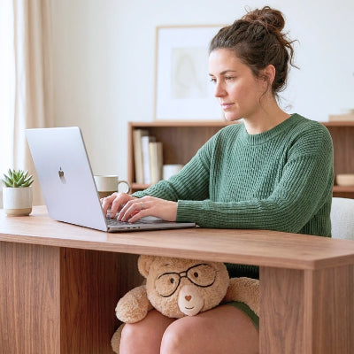 A woman in a green ribbed sweater sits at a wooden desk typing on a silver laptop. Percy the 5 lb comfort bear is wearing glasses sits under the desk, and a small potted plant and mug are on the desk beside her.