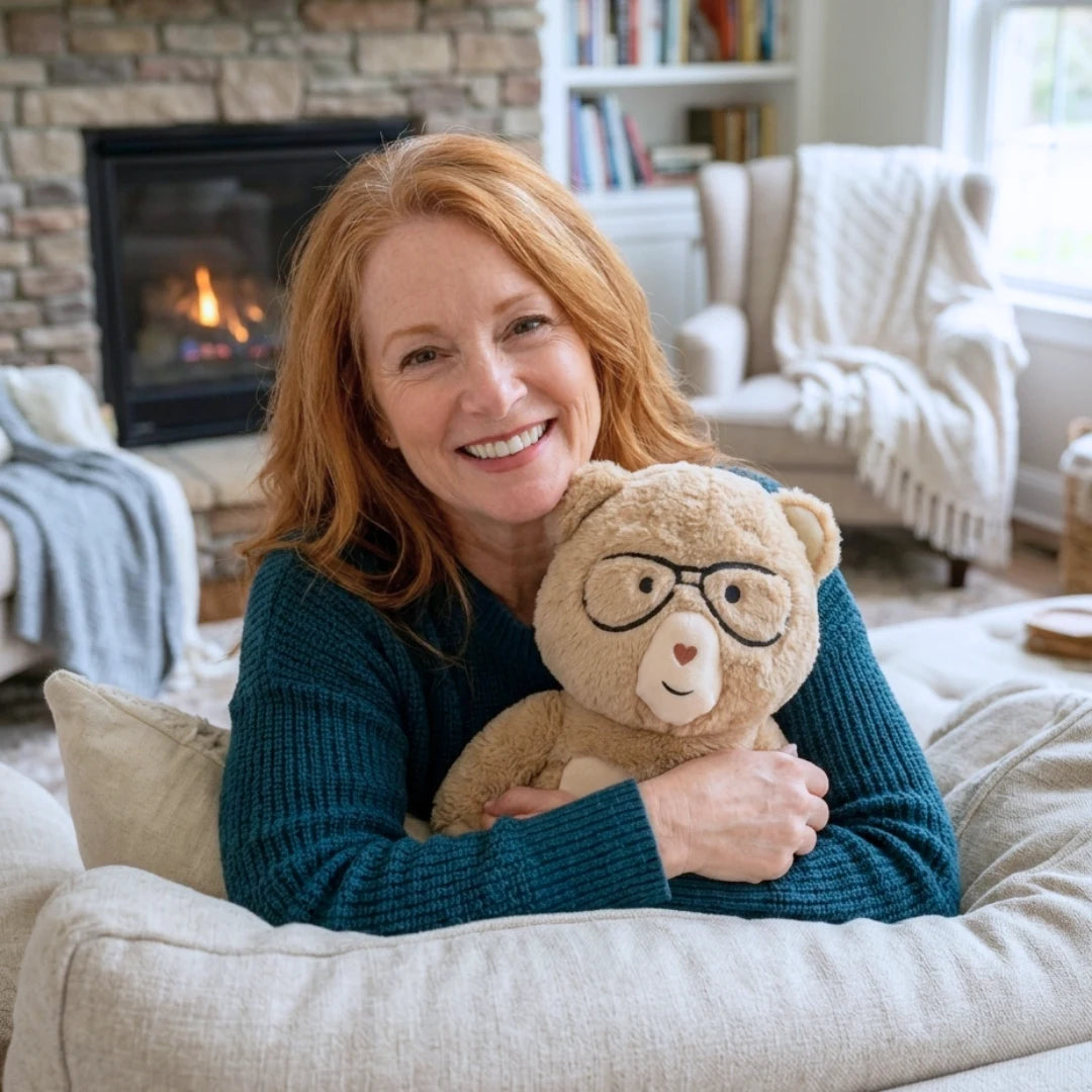 A smiling woman with red hair sits on a light beige couch in a cozy living room, holding Percy the 5 lb comfort bear. A fireplace, bookshelves, and upholstered chairs are visible in the background.