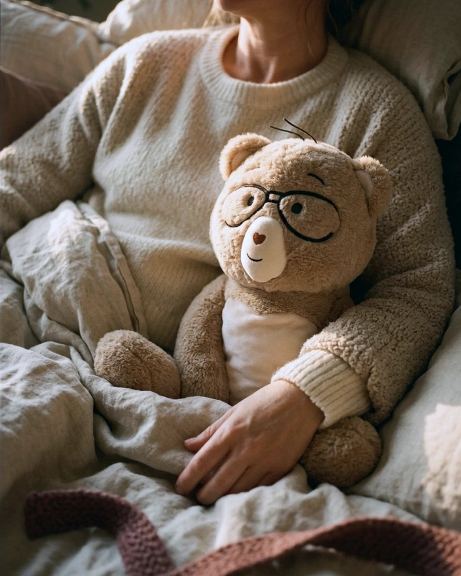 A cozy lifestyle shot of a person in a cream knit sweater holding a plush Oercy bear with black glasses and a white shirt, nestled under soft beige bedding.
