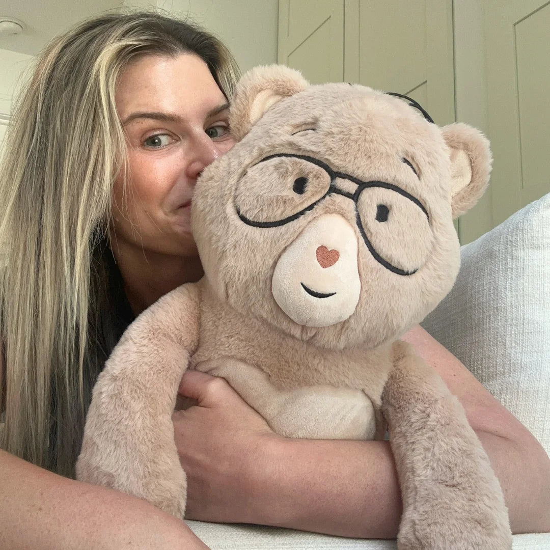Velle's founder, a woman with blonde hair holds Percy against her face. The teddy bear is light beige with black-framed glasses, a white snout, and a small red heart on its nose.