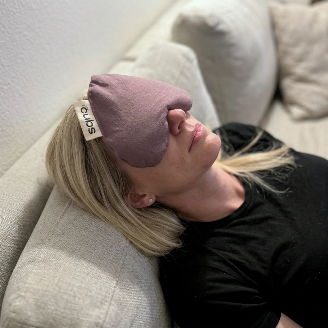 A woman reclining on a light beige couch with her eyes covered by a mauve heart shaped pouch. The mask has a small white fabric tab on the side with the brand name 'for the cubs' printed in black.