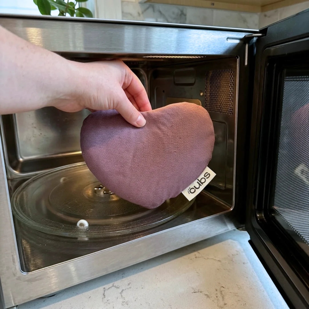 A hand holds a dusty rose heart-shaped thermal pouch inside a stainless steel microwave. The pouch has a small white tag with the brand name 'for the cubs' visible.