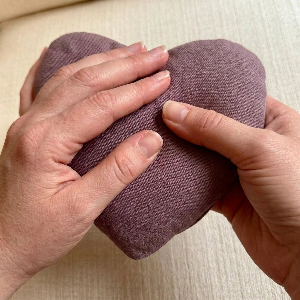 A close-up of a person's hands holding and gently pressing a heart-shaped, dusty mauve thermal heart pouch against a light beige textured background.