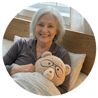 A smiling older woman with short gray hair lies in bed holding a beige teddy bear plush toy with glasses. The photo is framed in a circular border against a white background.