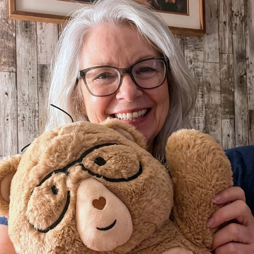 A smiling older woman with gray hair and glasses holds Percy the 5 lb. comfort bear with black glasses and a beige snout in front of a rustic wood-paneled wall.