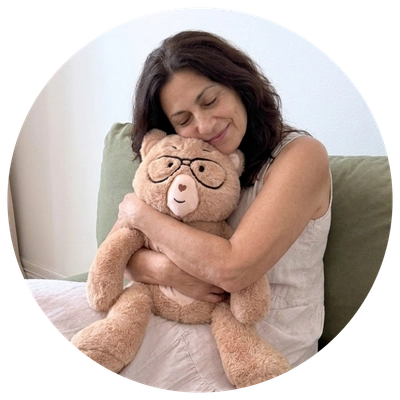A woman in a light-colored sleeveless dress is hugging a large beige teddy bear with black glasses, seated on a green couch against a light wall.