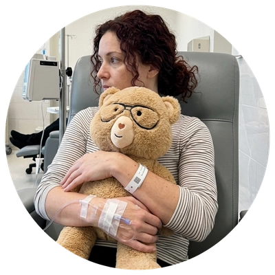 A woman with curly brown hair sits in a clinic chair holding a brown teddy bear with glasses. She has an IV in her arm and a wristband, with medical equipment visible in the background.