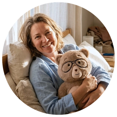 A smiling woman in a bedroom holds a brown teddy bear wearing round black glasses. The photo is framed in a circular border with a white background.