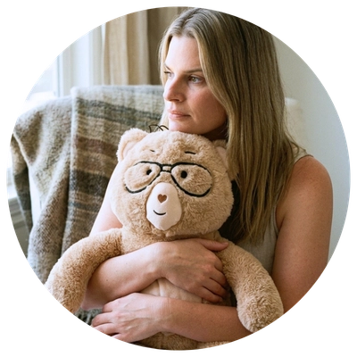 A woman with long blonde hair sits indoors holding a plush teddy bear with black-rimmed glasses. The photo is presented inside a circular frame against a light gray background.