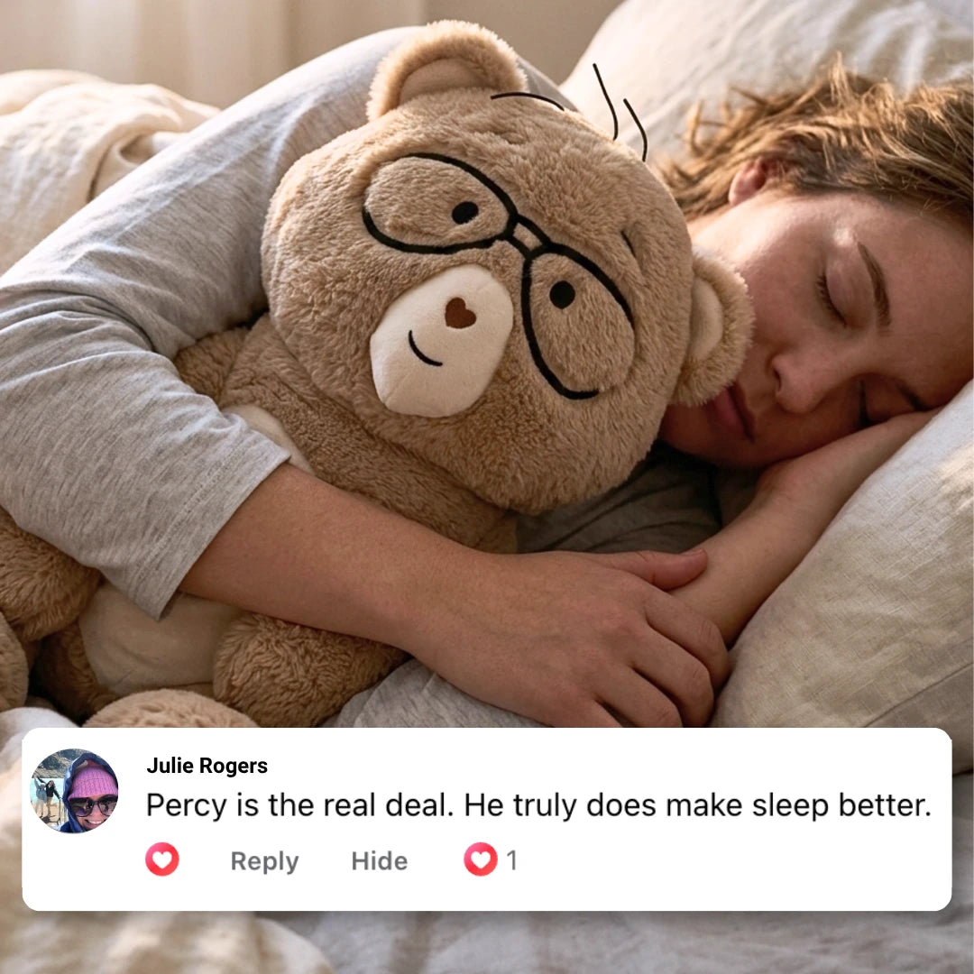 A person sleeping in bed with Percy the 5 lb comfort bear cuddled close. The bear is tan with black glasses and a cream snout.