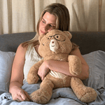 Woman holding a large teddy bear with glasses on a bed.