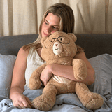 Woman holding a large teddy bear with glasses on a bed.