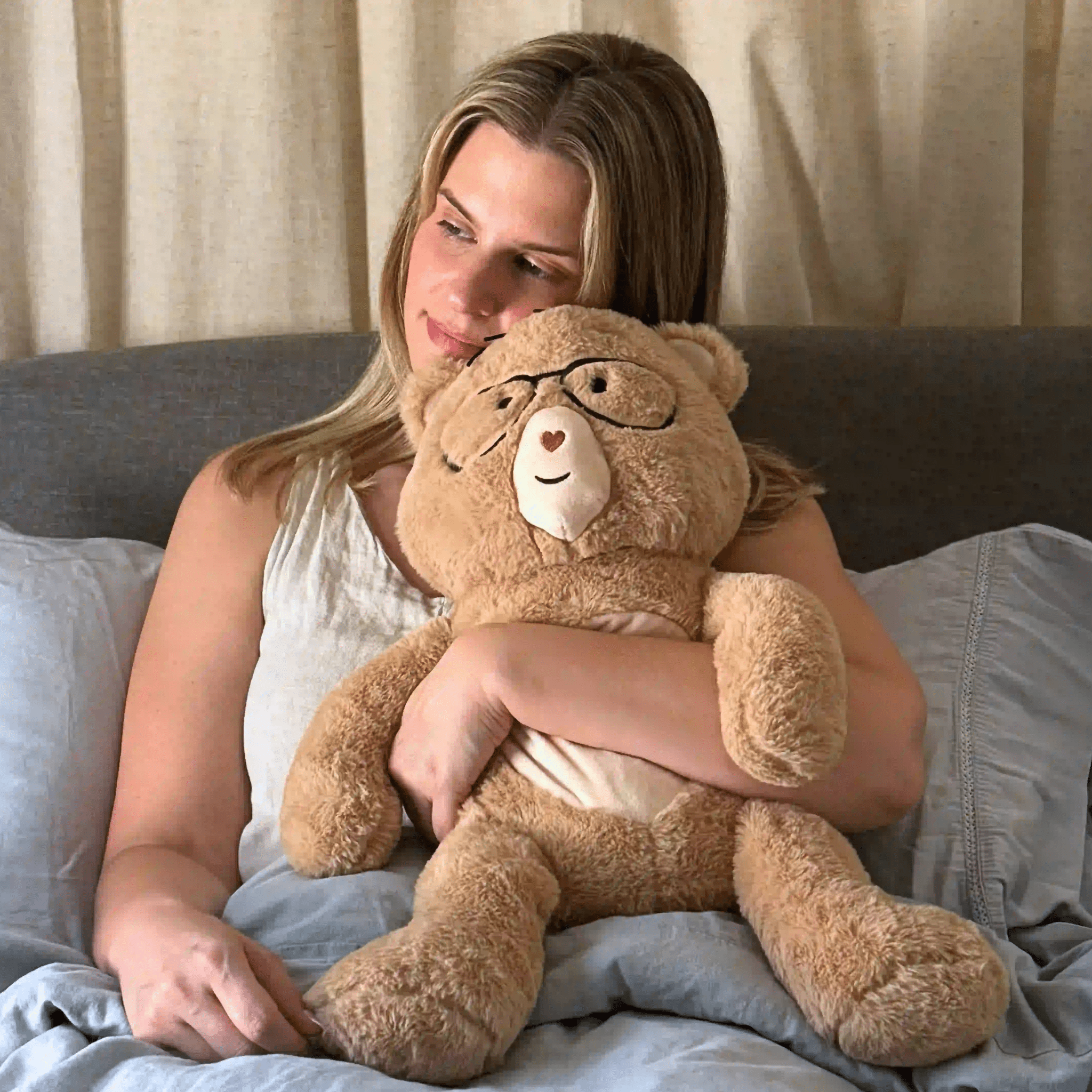 Woman holding a large teddy bear with glasses on a bed.