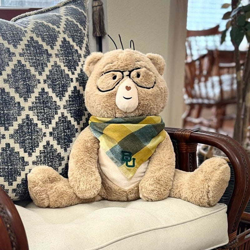 Percy bear with glasses, a cream face, and a yellow, green, and beige plaid bandana sits on a cream upholstered chair with a patterned blue and cream pillow behind it.