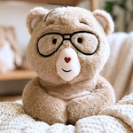 A beige plush teddy bear wearing black-rimmed glasses, sitting on a cream knit blanket in a softly blurred indoor setting.