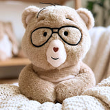 A beige plush teddy bear wearing black-rimmed glasses, sitting on a cream knit blanket in a softly blurred indoor setting.