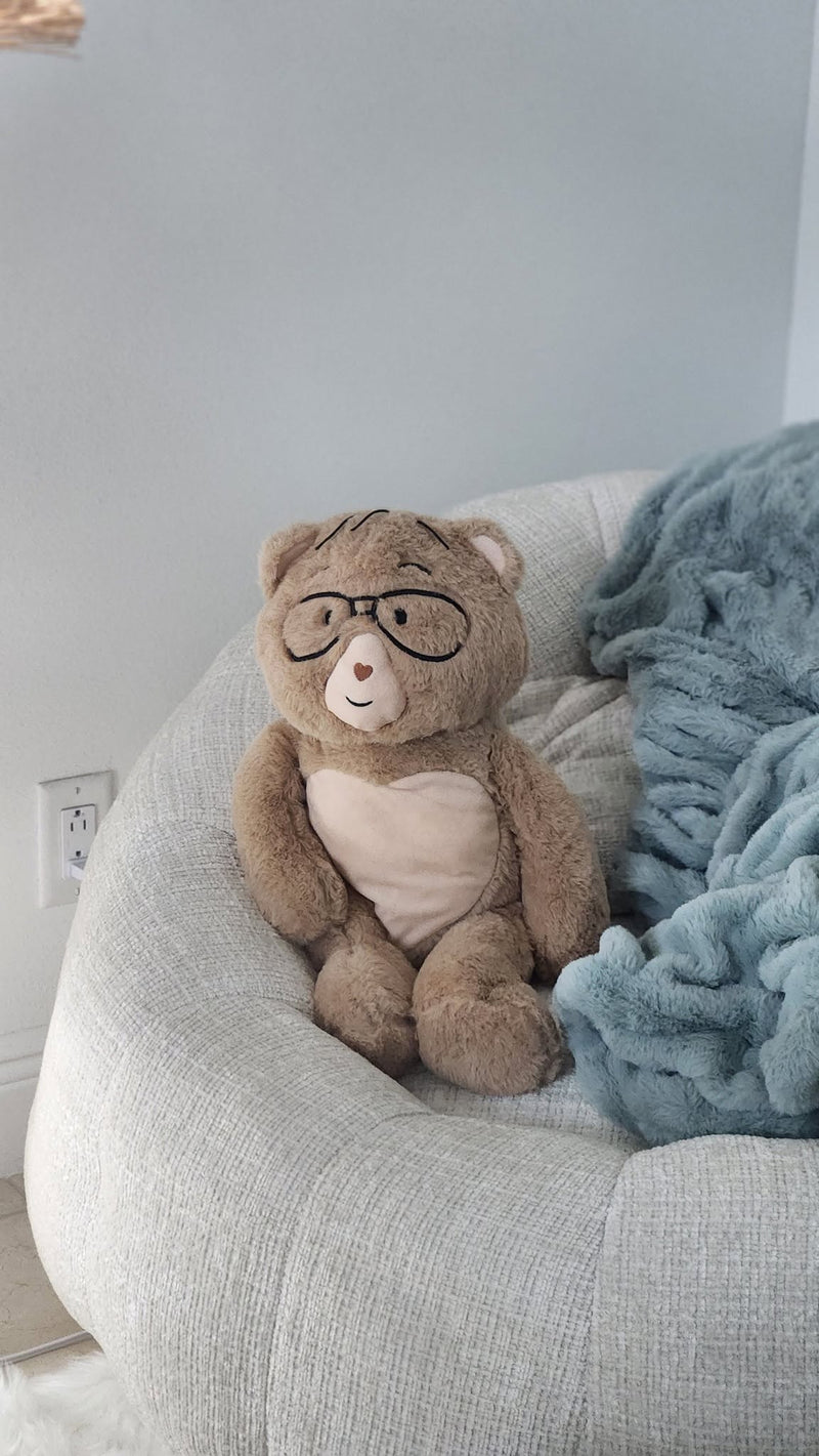 Percy the 5 lb. bear wearing round black glasses sits on a light gray fabric chair next to a chunky blue throw blanket in a softly lit indoor setting.