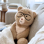A plush teddy bear toy with black-rimmed glasses, a cream snout, and a small red heart on its nose, sitting on a light gray couch in a cozy living room setting.