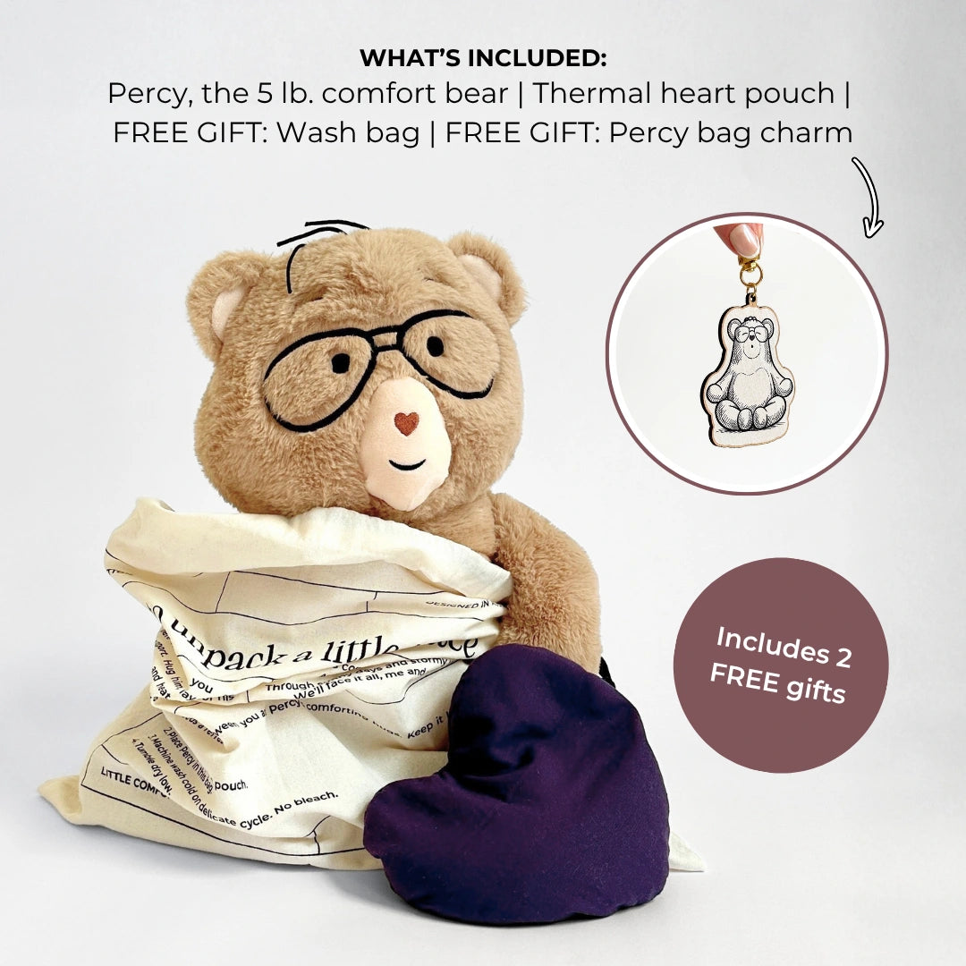 A teddy bear comfort bear wearing glasses, wrapped in a cream bag, with a dark purple heart-shaped pouch beside it. Text at the top lists included items: the bear, a thermal heart pouch, a wash bag, and free gift bag charms.