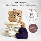A teddy bear comfort bear wearing glasses, wrapped in a cream bag, with a dark purple heart-shaped pouch beside it. Text at the top lists included items: the bear, a thermal heart pouch, a wash bag, and free gift bag charms.